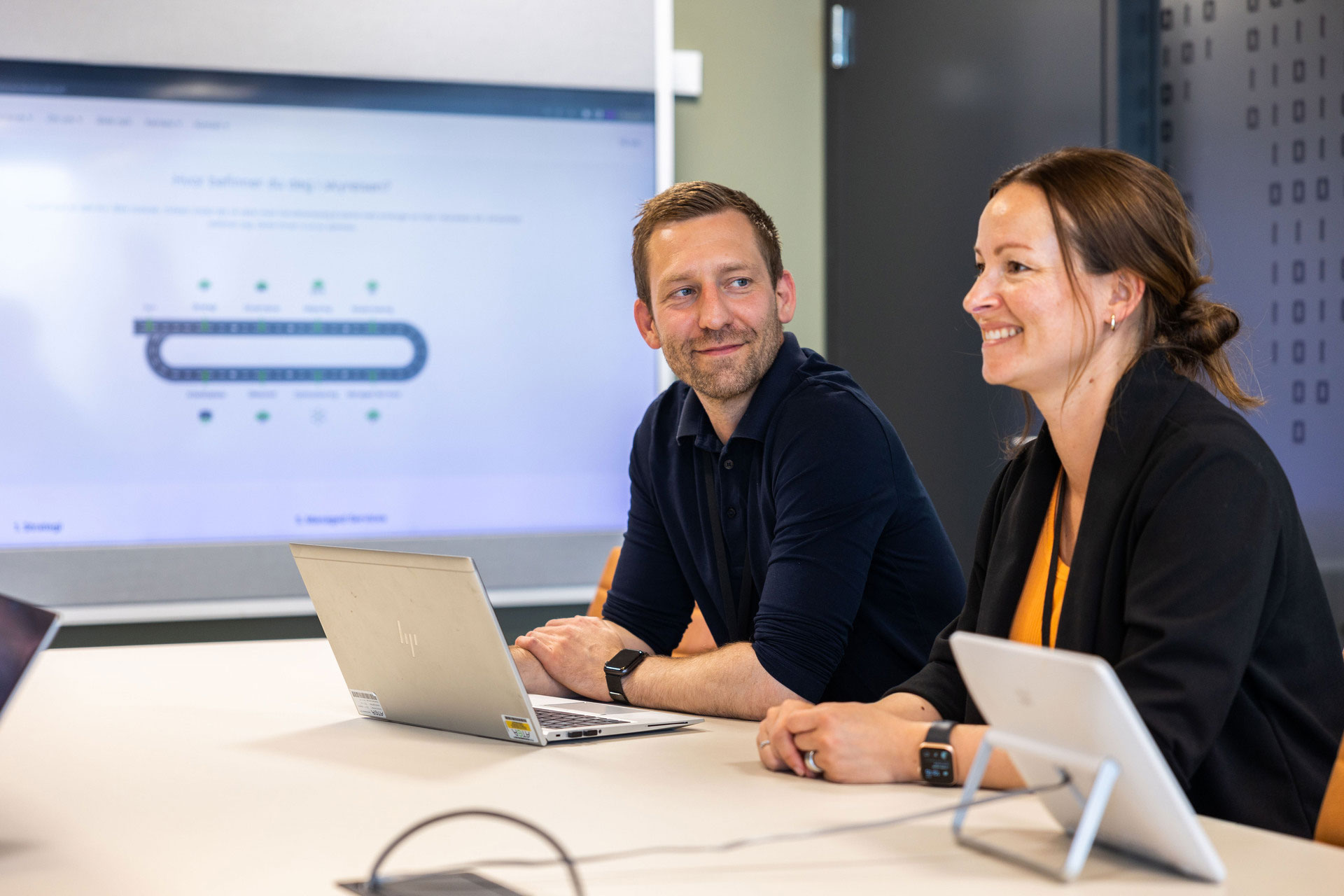 A Man And A Woman Sitting In Meetingroom With Big Screen In The Back And PC On Table